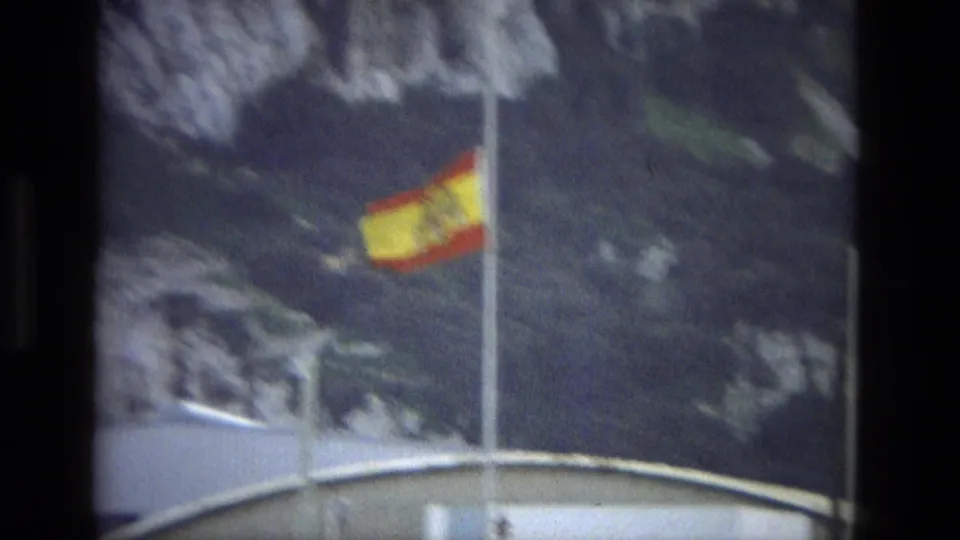 The Spanish flag is seen closer in the frame, waving beside a pole with rocky terrain in the background.