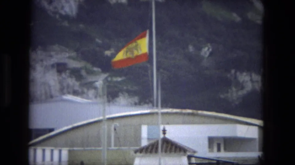 A Spanish flag on a tall pole rises above a pale roofline and rocky landscape in the 1978 Cordoba clip.