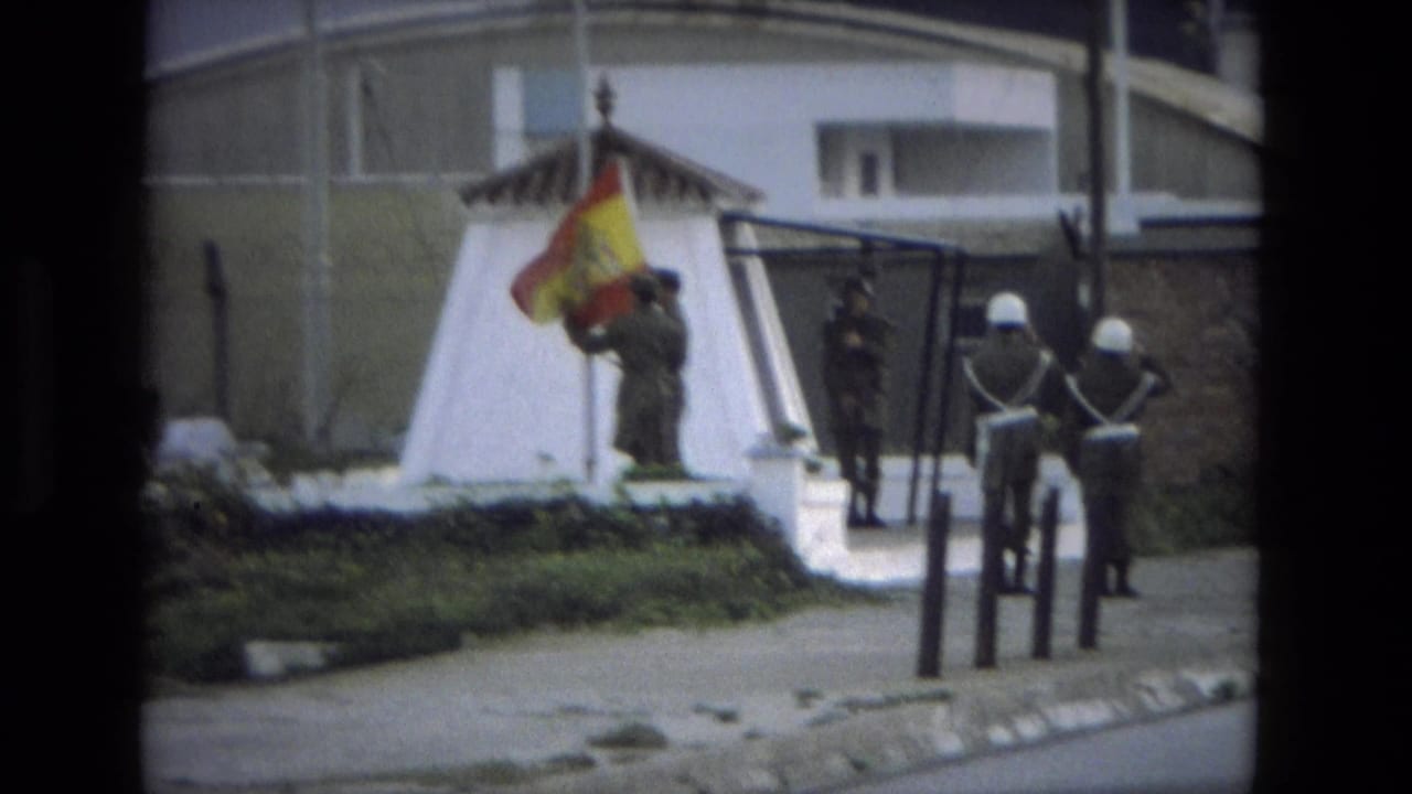 Soldiers stand near a Spanish flag at a white roadside monument or flag base.