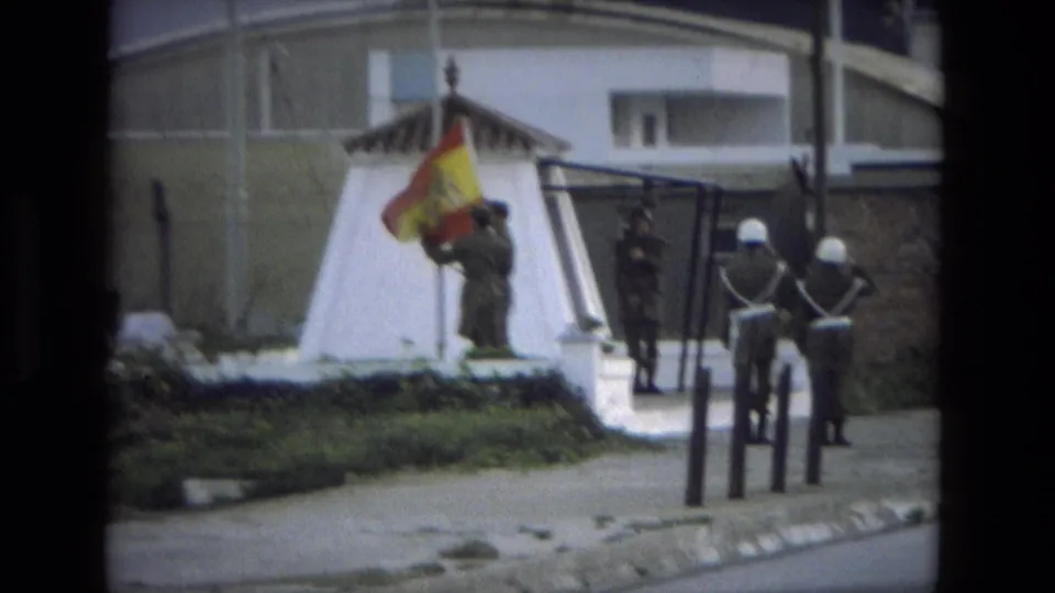 1978 Spain 8mm frame showing uniformed soldiers by a Spanish flag beside a white monument or flag base and street paving.