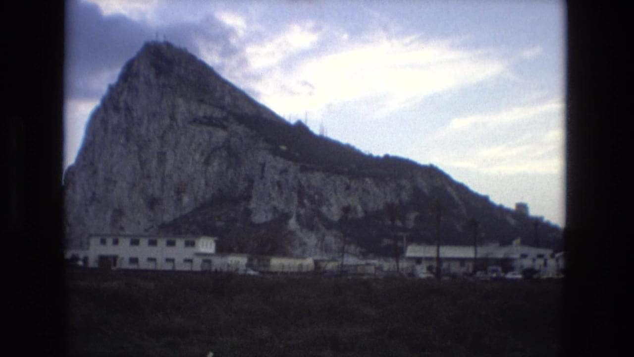 1978 Spain 8mm view of a large rocky landmark with low buildings in the foreground and clouded sky above.