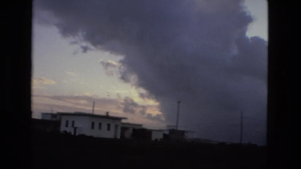 1978 8mm frame of low buildings, utility poles, and a large cloud bank at dusk in the Spain location supplied by metadata.