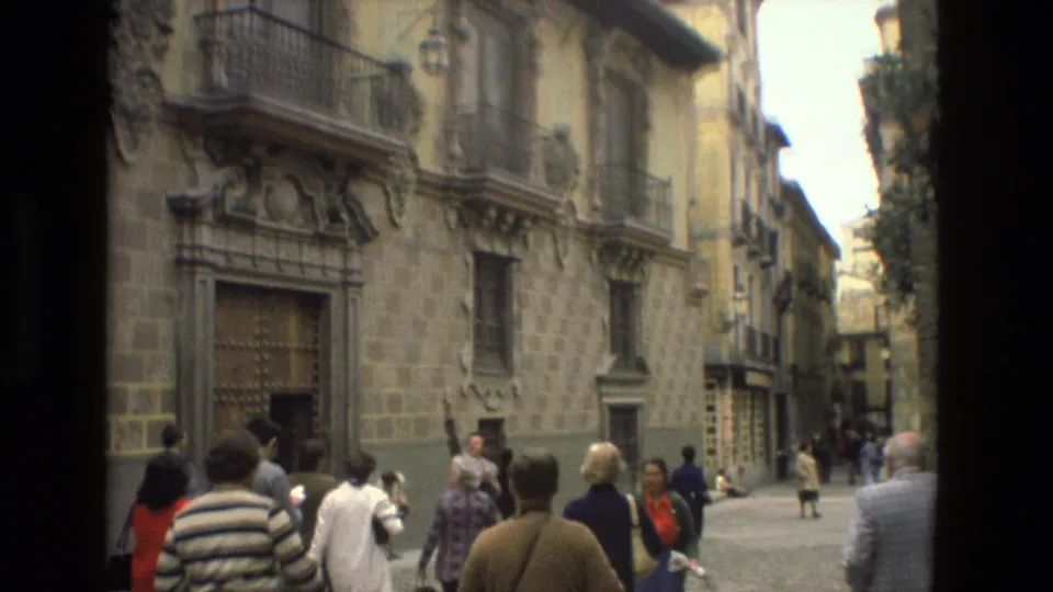 Tourists in 1970s casual clothing walk through a Córdoba street bordered by historic stone facades and ornate balconies.