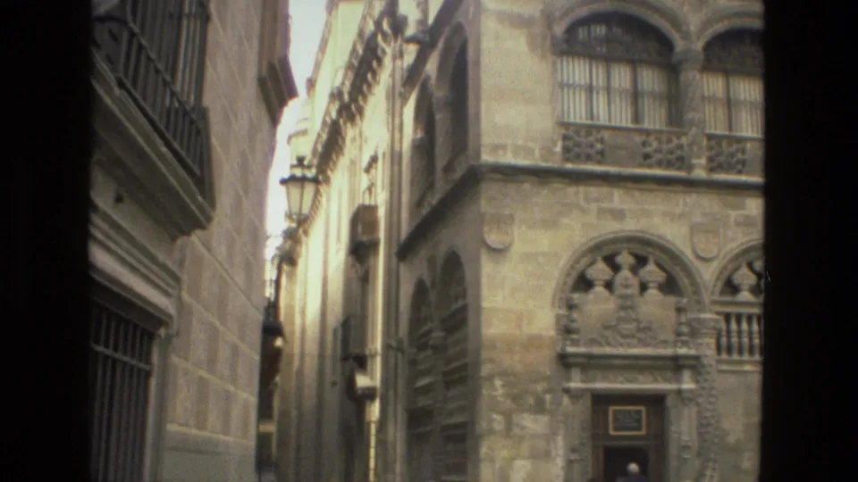 Handheld 8mm view of a Córdoba stone facade with arched windows, barred openings, a doorway, and a narrow lane at left.