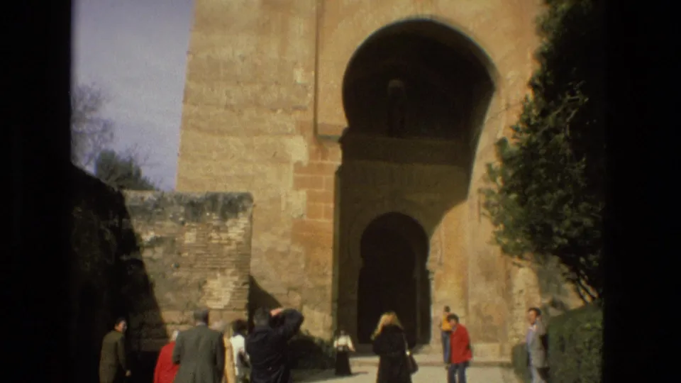 Visitors in 1970s clothing gather near a historic arched stone entrance in Córdoba, Spain, filmed on 8mm in 1978.