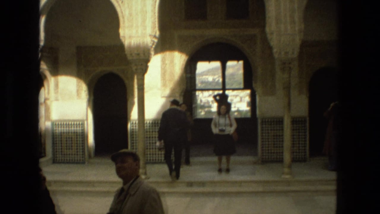 Tourists stand and walk beneath arched columns in a sunlit courtyard interior.