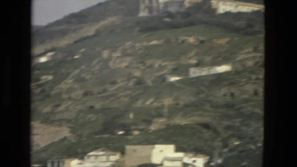 Soft 8mm frame from Córdoba, Spain, showing scattered white structures, hillside tracks, and open green-brown terrain.