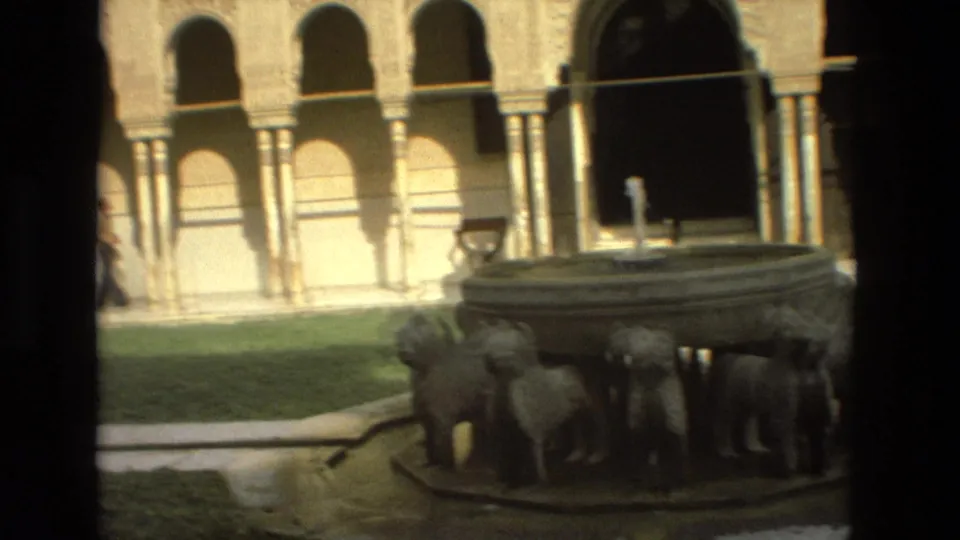 1978 8mm frame from Córdoba showing a courtyard fountain with lion sculptures, grass, columns, and arched openings behind it.