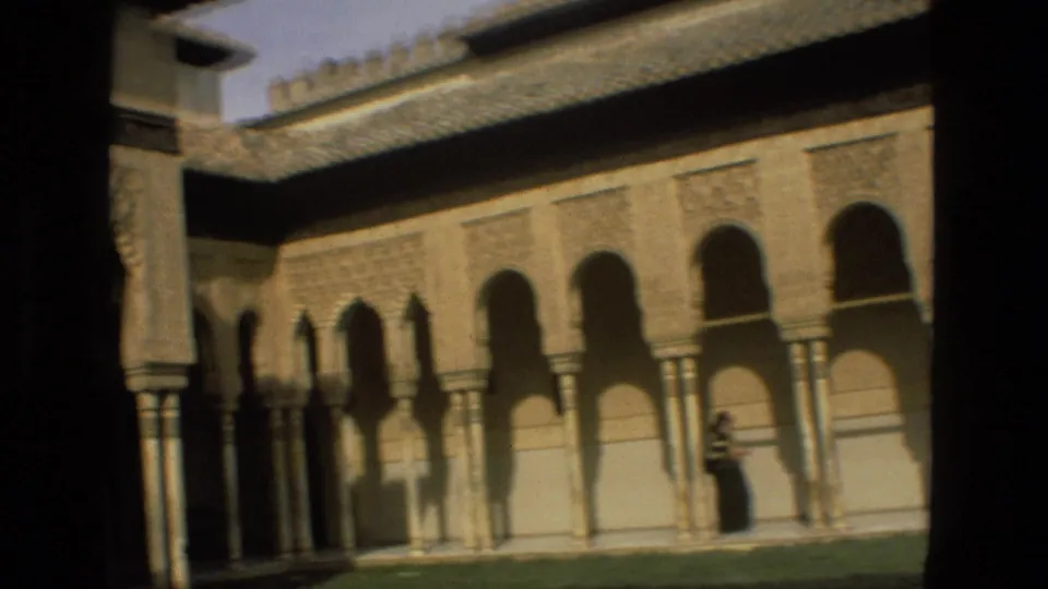 Córdoba, Spain, 1978 home-movie frame showing a long Moorish-style colonnade, tiled roofline, grass, shadows, and a distant visitor.