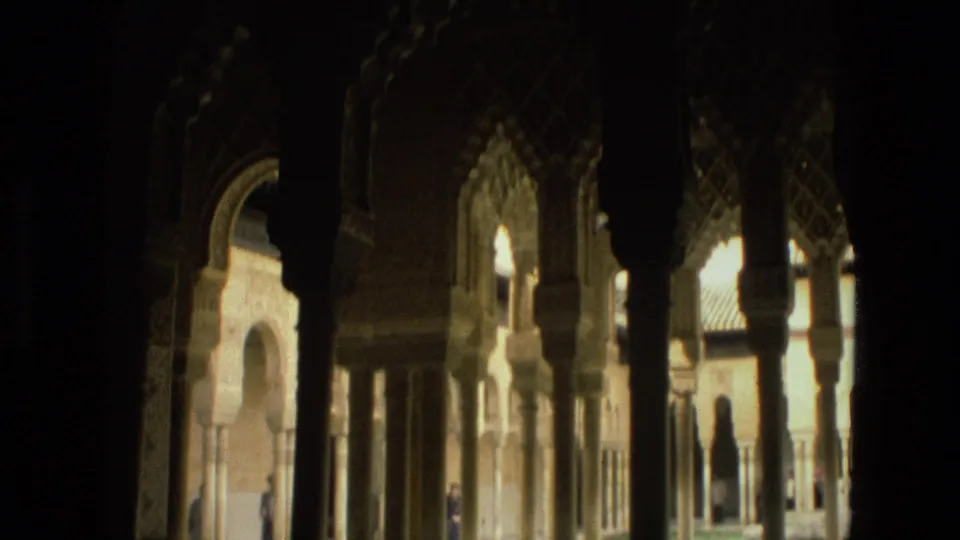 Soft 8mm frame of a Cordoba courtyard, with dark foreground columns, arched openings, and sunlit garden architecture beyond.