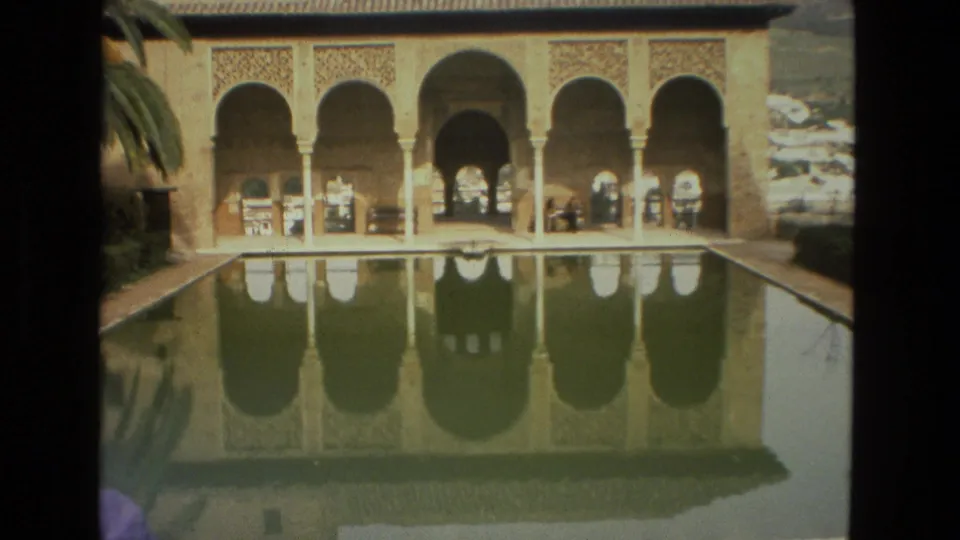 A visitor-shot 8mm view of Córdoba arches mirrored in still water, with people visible near the arcade.