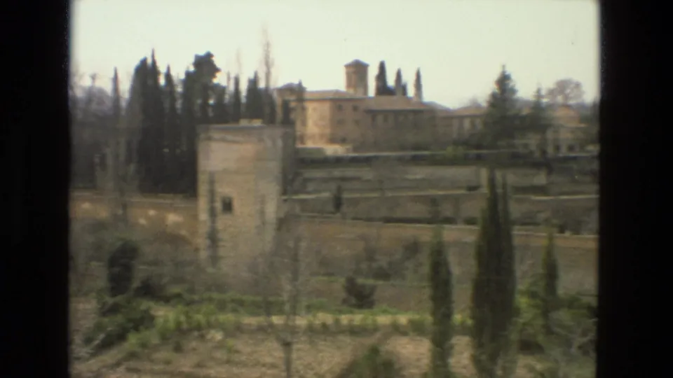 1978 Cordoba 8mm frame with formal garden walls and cypress trees in front of a larger historic stone building and tower.