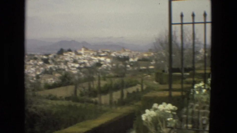 Córdoba city panorama on 8mm film with white buildings, hazy hills, clipped garden hedges, flowers, and iron railings.