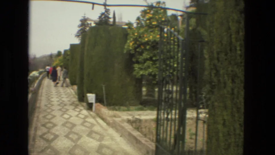 Visitors walk along a patterned stone path bordered by clipped hedges and flowering plants in a Cordoba garden filmed on 8mm in 1978.