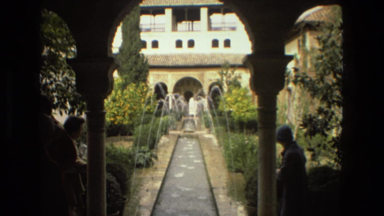 Shaded arches frame a courtyard with fountain jets, a long water channel, greenery, and two visitors in silhouette.