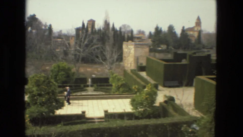 Wide 1978 Córdoba garden view with terraces, clipped hedges, stone walls, distant towers, and one visible visitor.