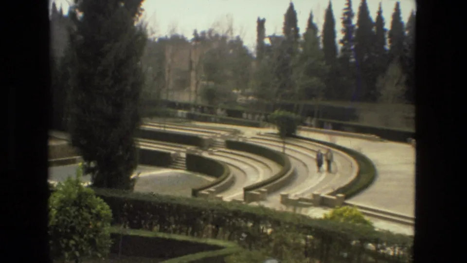 Vintage 8mm Córdoba garden view showing curved stone steps, hedges, tall trees, and two distant visitors crossing the walkway.