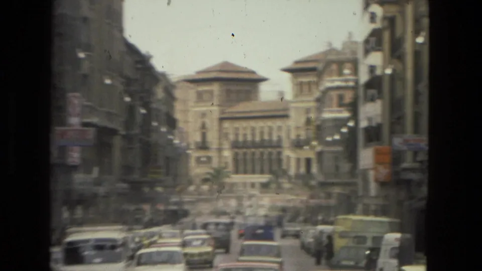 Blurred 1978 Córdoba street frame showing cars, pedestrians, dense facades, and a large arched-window building beyond the traffic.
