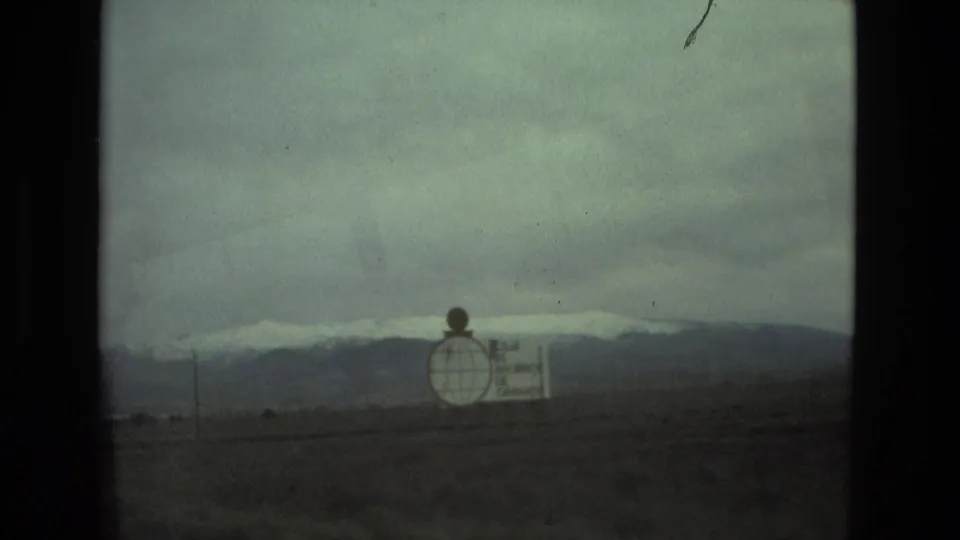 Wide landscape view from the 1978 travel footage, showing a roadside sign, dark ground, overcast sky, and distant snow-capped mountains.