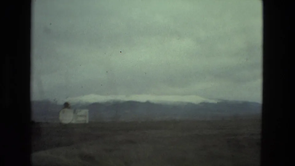 Roadside view in Spain with a small sign in the foreground, open land, heavy clouds, and a pale snow line on distant mountains.