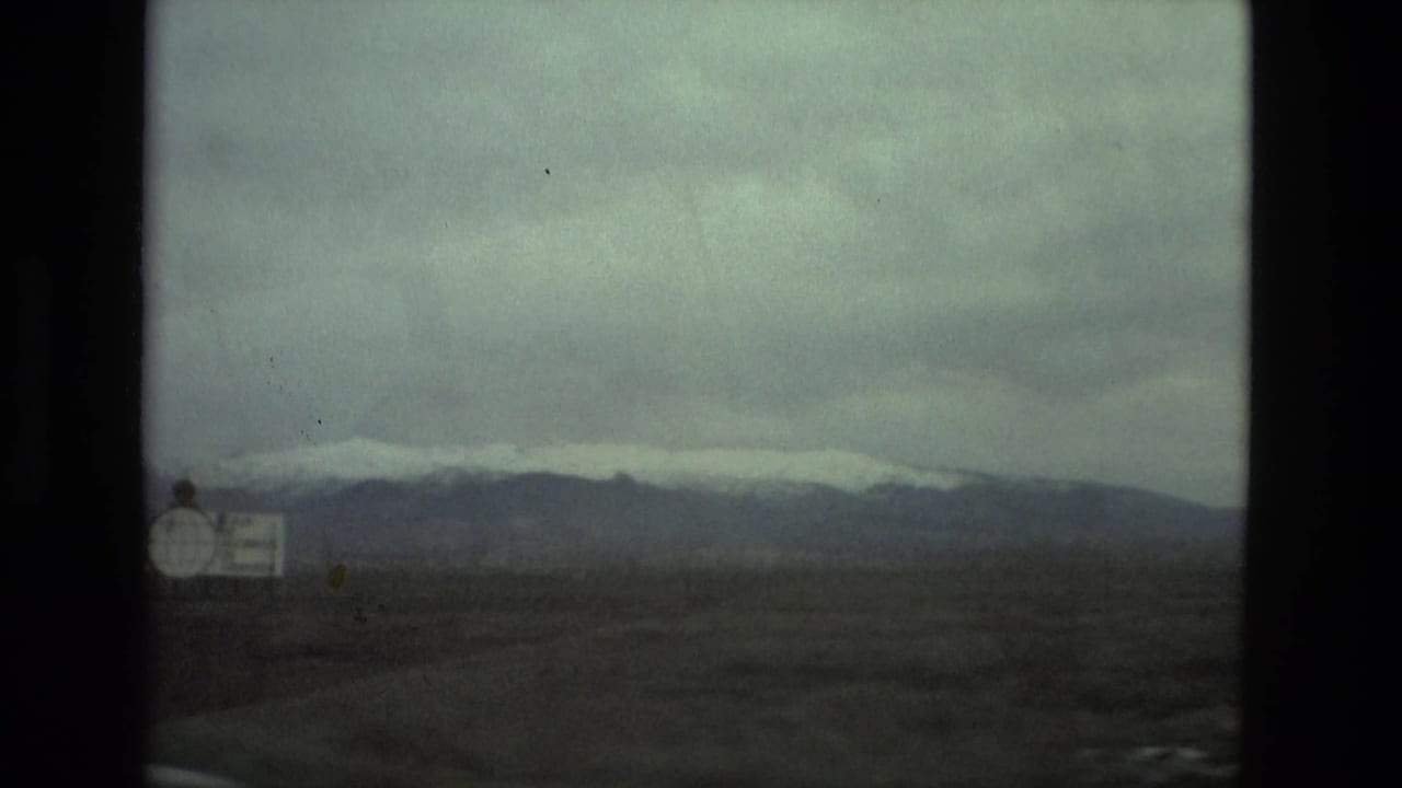 Roadside sign at the edge of a dry plain with distant snowy mountains under a gray sky.