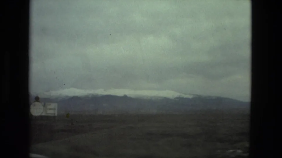 1978 8mm roadside view near Seville, Spain, showing a white sign, open plain, and snow-capped mountains through a dark vehicle window frame.