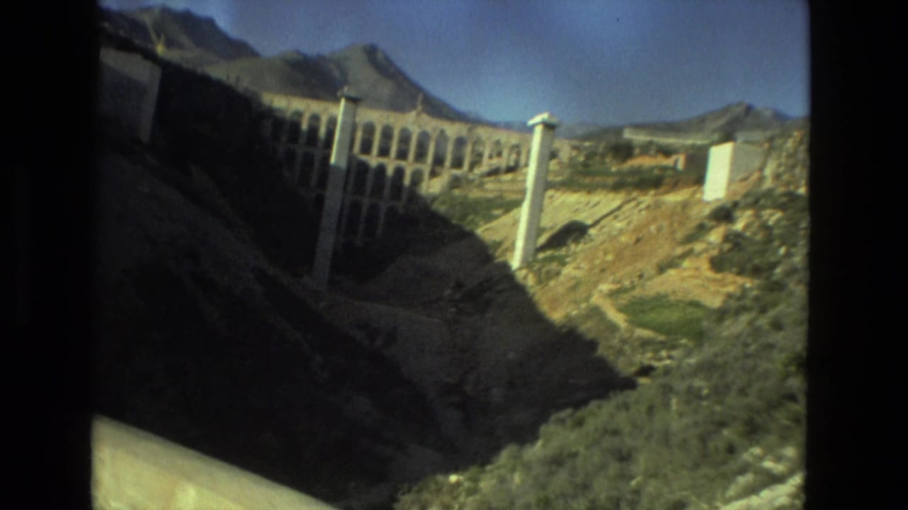 1978 8mm travel frame from Seville showing a sunlit valley, tall pale supports, dry hillside, and distant mountains.