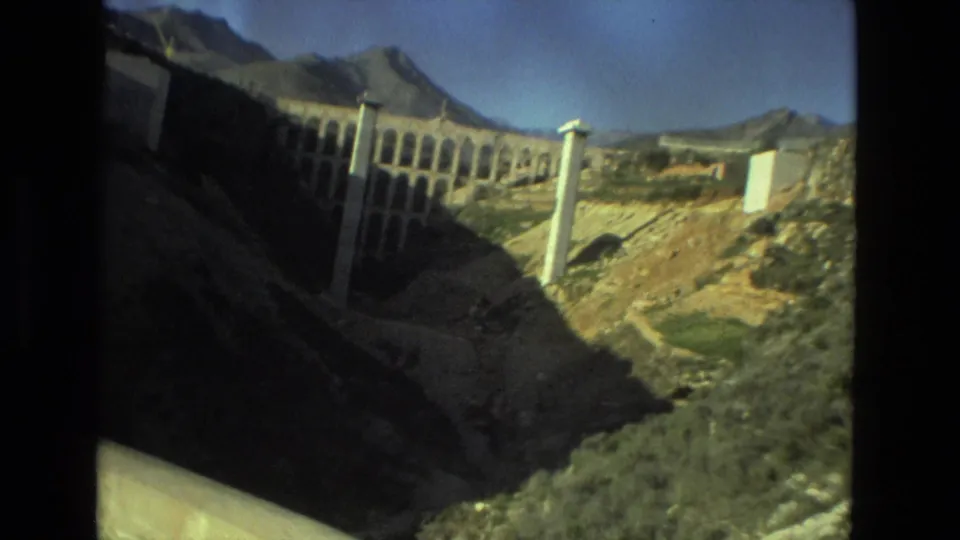 1978 8mm travel frame from Seville showing a sunlit valley, tall pale supports, dry hillside, and distant mountains.
