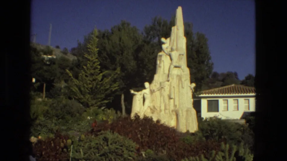 1978 Seville home-movie frame showing a white climbing-figure monument in landscaped greenery beside a small tiled-roof building.