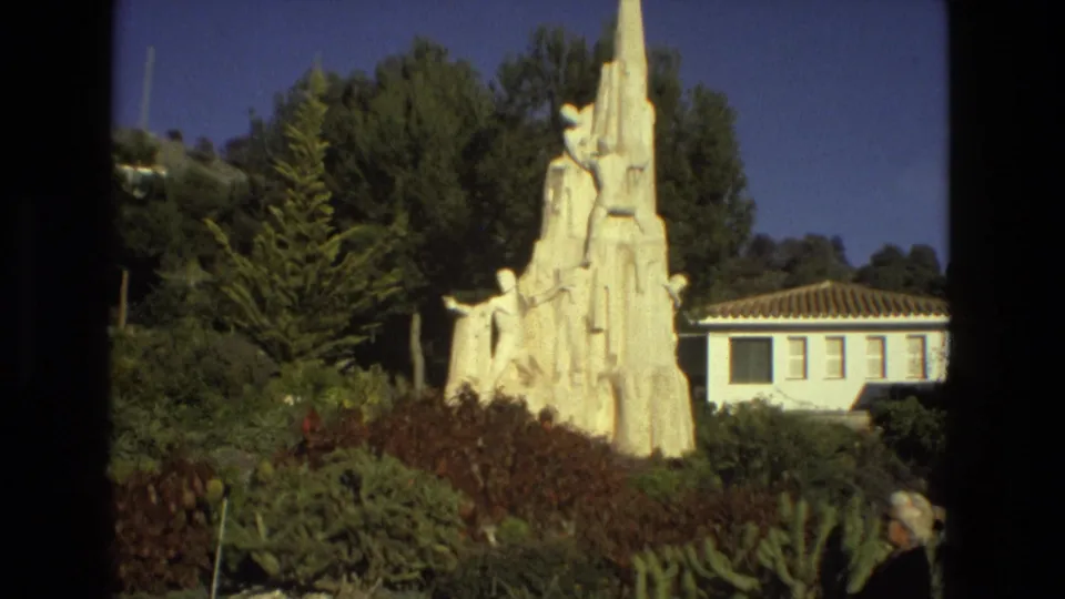1978 Seville 8mm frame with the white grouped-figure monument centered against shrubs, hillside trees, and a tiled-roof building.