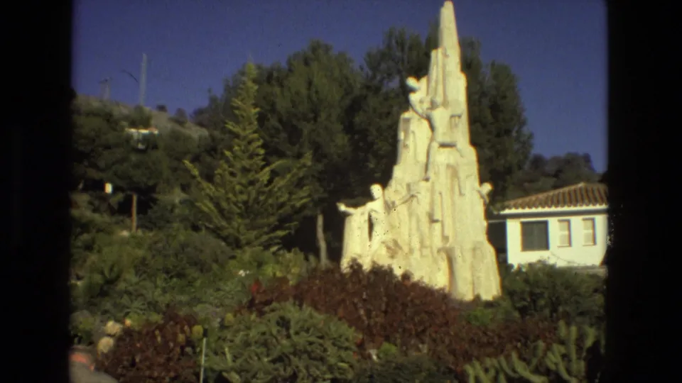 Final 1978 Seville 8mm frame holding on the white climbing-figure monument amid greenery, hillside, and a tiled-roof building.
