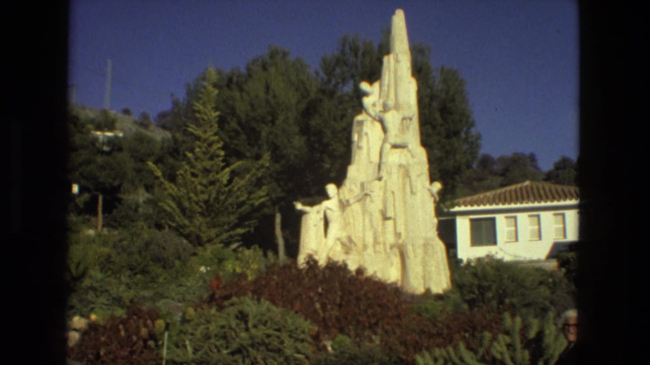 White sculptural monument rises above plants in a sunny garden with a tiled-roof building nearby.