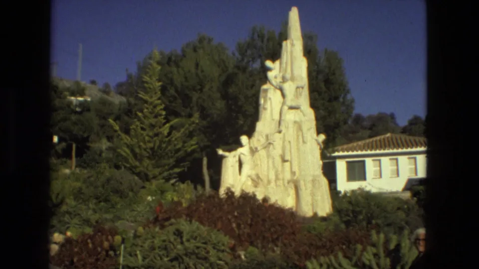 Seville, Spain, 1978 home-movie frame showing a white monument among garden plantings beside a low building with a tiled roof.