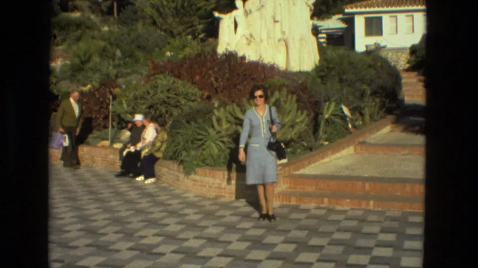 Seville, Spain, 1978 frame with a woman in a light blue suit and sunglasses walking past seated visitors in a sunlit garden.