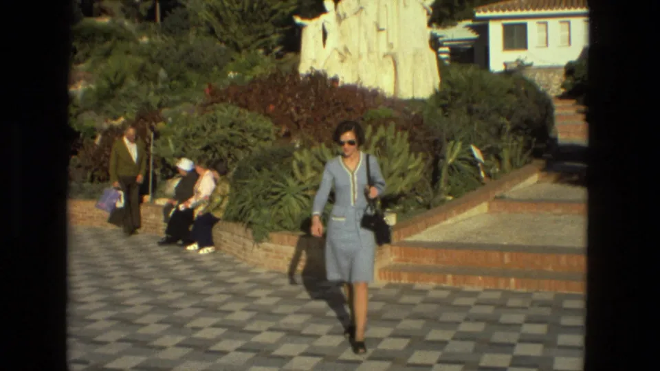 Seville, Spain, 1978 frame of a woman in sunglasses walking toward the camera on checked paving near a brick garden wall.
