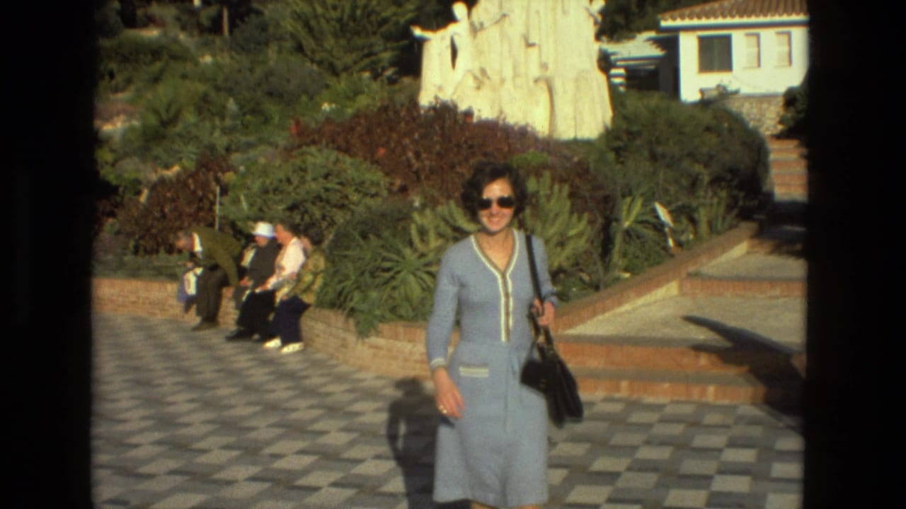 Woman in a light blue dress walks across checkered paving while visitors sit nearby.