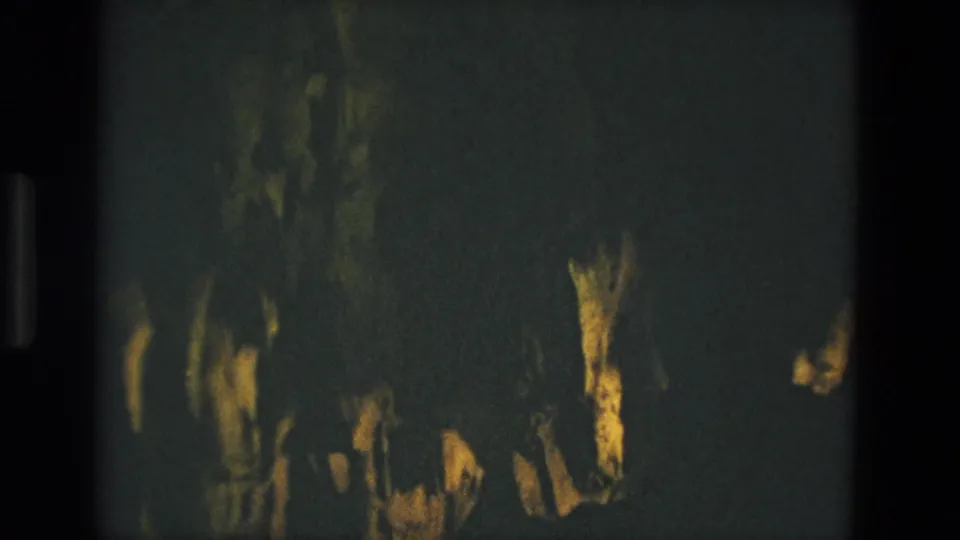 Final 8mm frame from the Seville cave clip, showing uneven rocky surfaces in deep shadow with yellow-green illumination.
