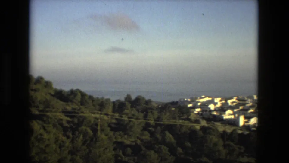 1978 8mm travel frame of a white hillside town above trees, with open water and a hazy sky beyond.
