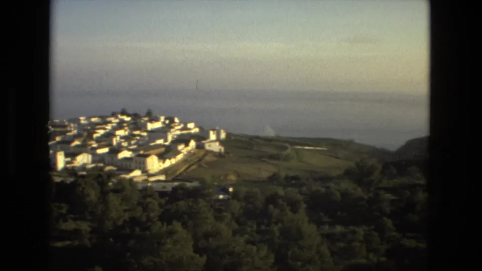 1978 8mm frame with the white town centered above trees and fields, with a hazy sea view in the background.