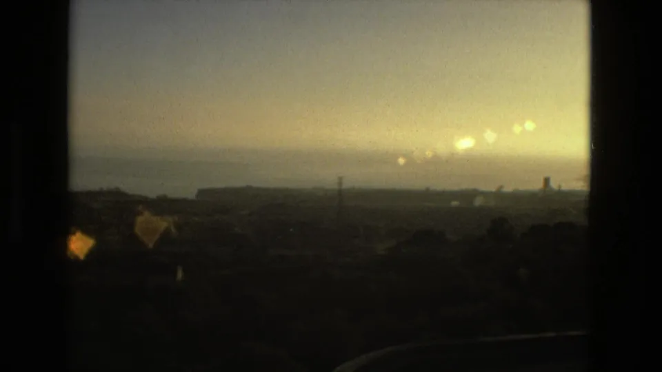 1978 Spanish coastal view with dark foreground hills, distant sea, haze, and warm lens flare on 8mm film.