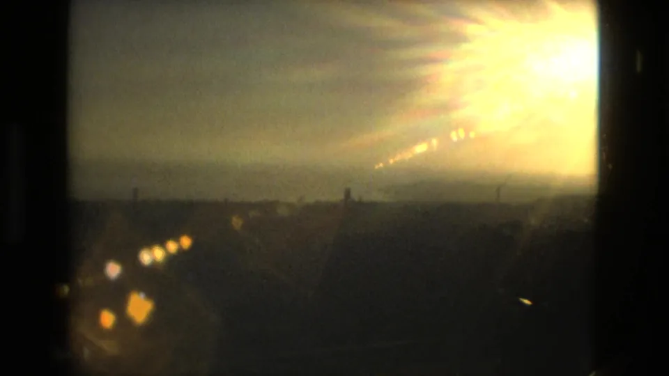 A warm 8mm view of intense sun flare above a distant coastal-looking landscape, framed by dark film edges.