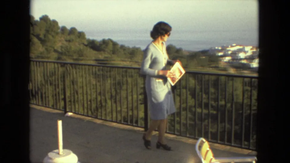 In metadata-labeled 1978 Spain, a woman in a light blue dress and sunglasses walks along a balcony with trees, white buildings, and water beyond.