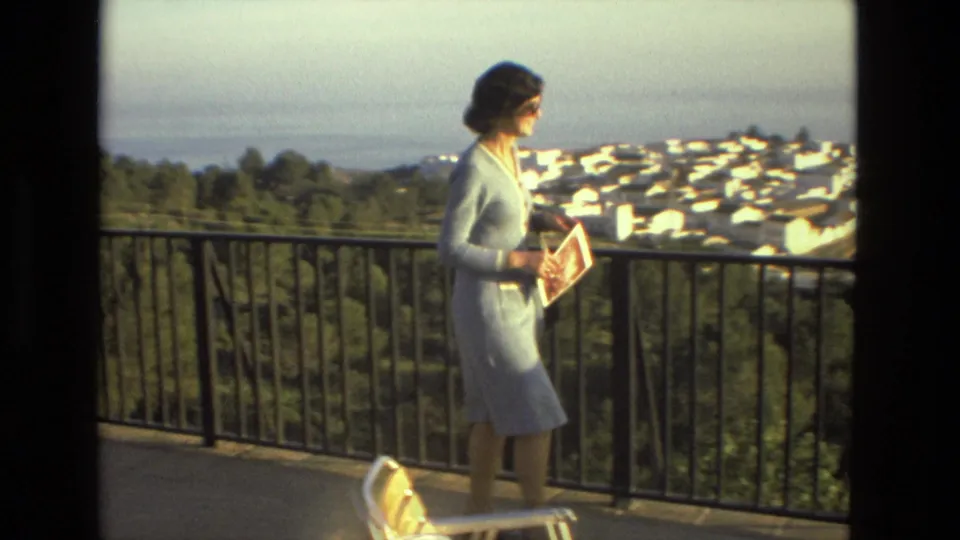 A woman in a light blue dress holds a booklet by a balcony railing, with a hillside, white buildings, and distant water behind her.
