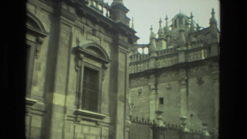1978 8mm view in Seville showing a close stone corner, barred window, iron fence, balustrade, and ornate upper facade.