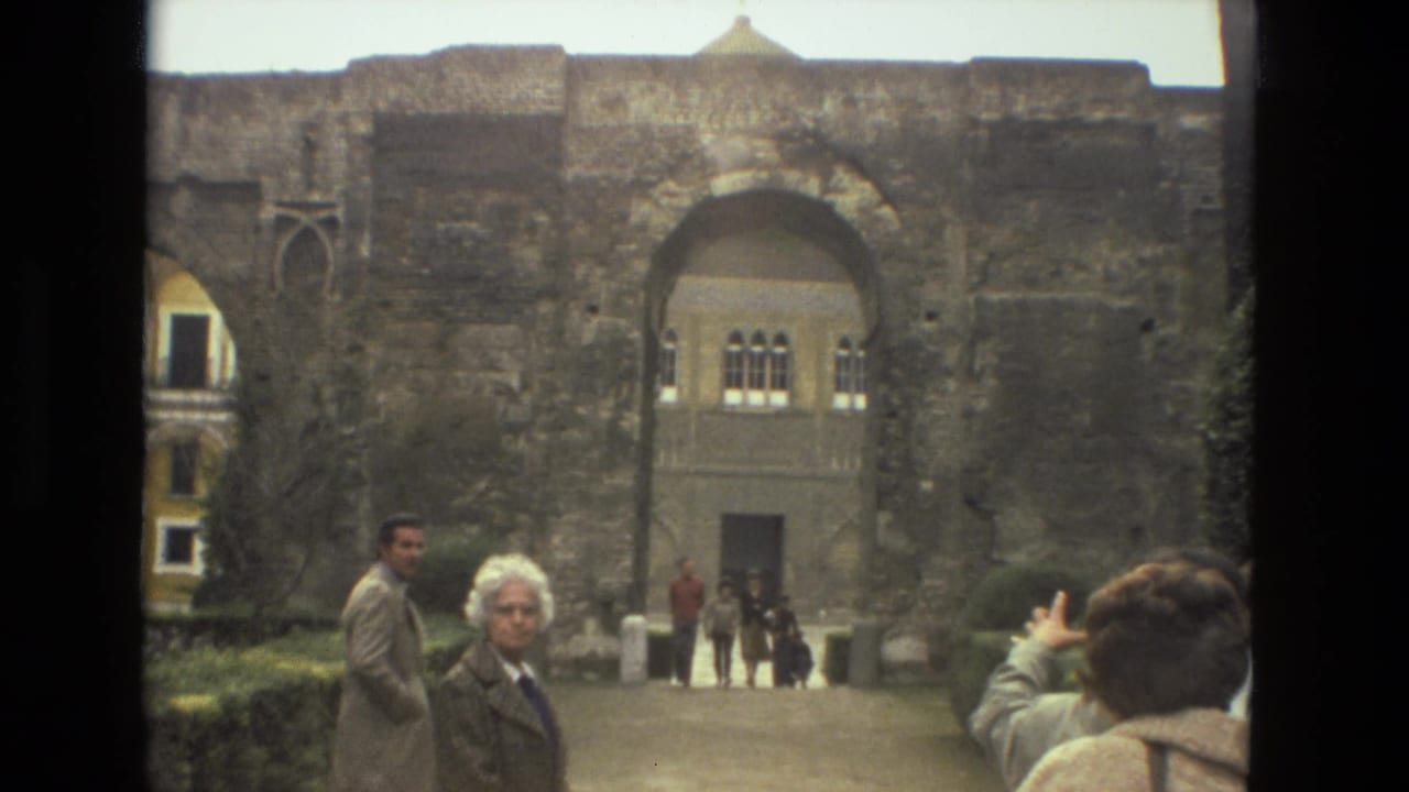 Seville 1978 frame of tourists in a courtyard facing a large stone arch with an ornate building visible beyond it.