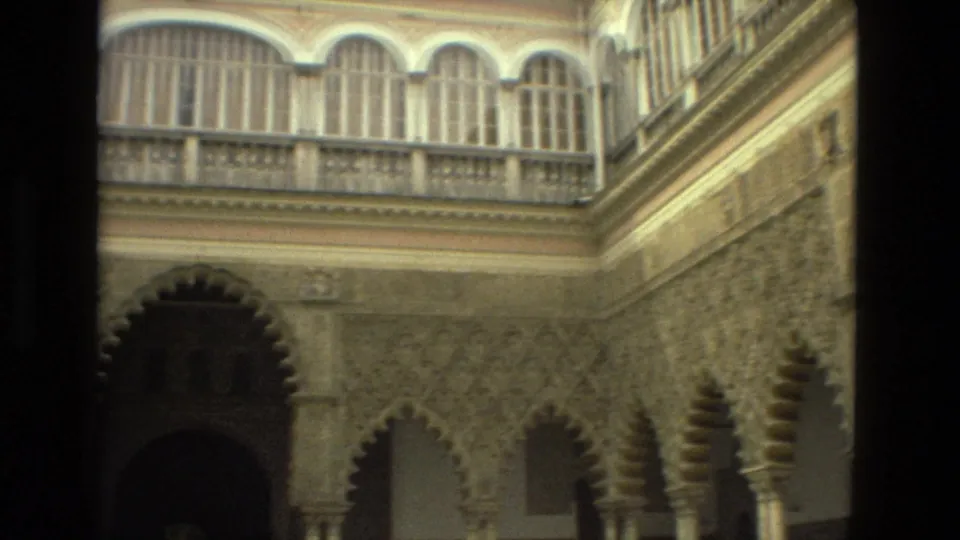 A soft 8mm frame shows lower scalloped arches, slender columns, and an upper gallery of arched windows in Seville, Spain.