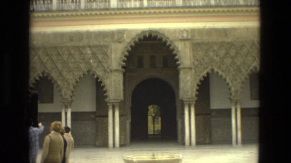 1978 8mm Seville courtyard scene with a central stone fountain, ornate arches, and visitors walking at the edge of frame.