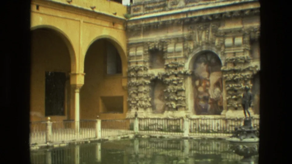 1978 Seville courtyard view with yellow arcades, reflective pool, ornate wall relief, small fountain, and one distant visitor.