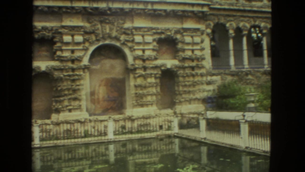 Ornate stone garden building reflected in a pool behind an iron fence.