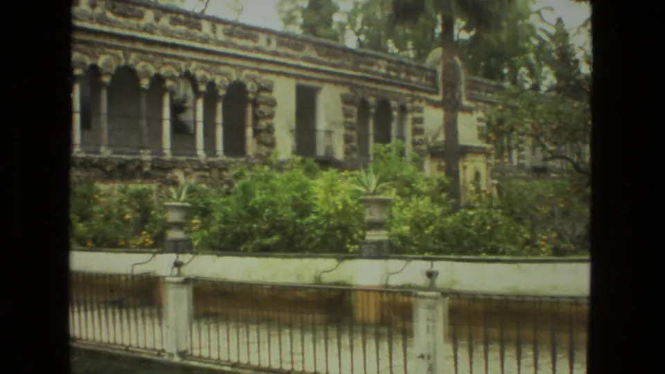1978 Seville home-movie frame showing garden shrubs, iron railing, water, and an ornate stone building.